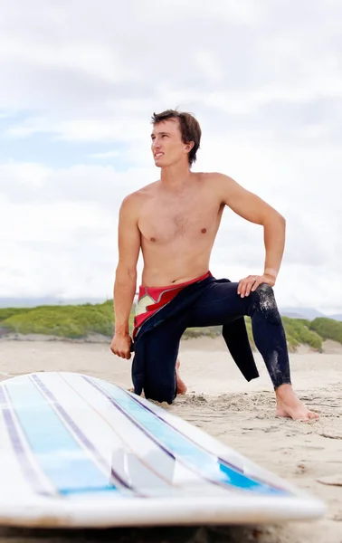 Vorbereitung auf die Wellen. Ein junger Surfer macht Stretches am Strand neben seinem Brett. — Stockfoto