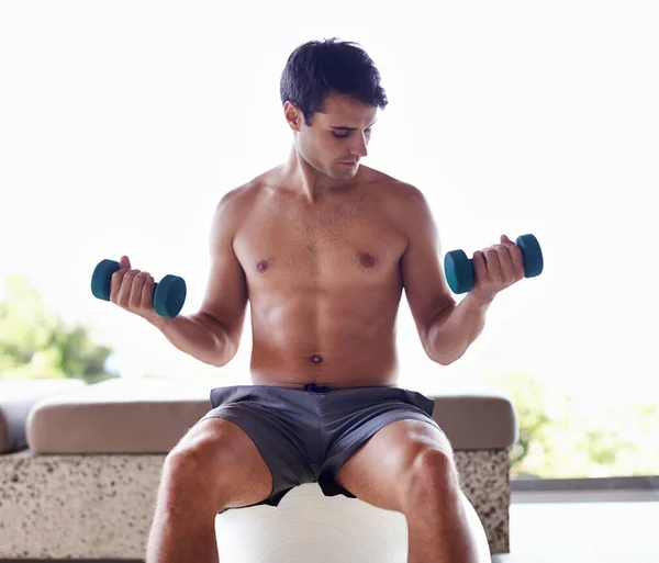 Υγιής πρωινή ρουτίνα. Ένας μυώδης νέος σηκώνει αλτήρες ενώ κάθεται σε μια μπάλα γυμναστικής.. — Φωτογραφία Αρχείου