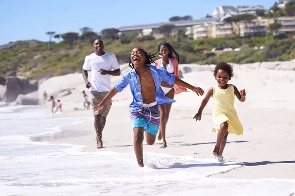 Nada é melhor do que umas férias em família. Uma jovem família africana feliz correndo energeticamente ao longo da praia. — Fotografia de Stock