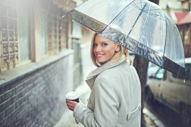 Warming up with a coffee. Rearview portrait of an attractive young woman walking in the rain with an umbrella and a coffee. clipart
