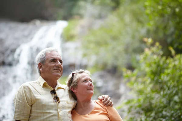 Vilken vacker dag att vara ute i naturen. Ett kärleksfullt äldre par som står mot bakgrund av ett pittoreskt vattenfall. — Stockfoto