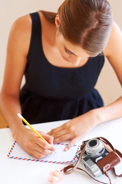 Piszę do domu. Młoda kobieta pisząca adres na kopercie z kamerą na biurku. — Zdjęcie stockowe