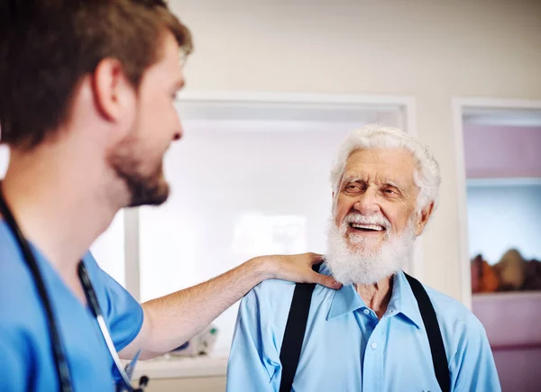 Siempre está feliz de ayudar a los ancianos. El disparo de un joven doctor poniendo su mano en un hombro de un hombre mayor en comodidad en una clínica. — Foto de Stock