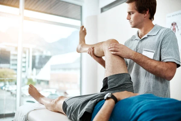 Te arreglaré en poco tiempo. Inyección de un joven fisioterapeuta masculino ayudando a un cliente con ejercicios de piernas que yace en una cama. —  Fotos de Stock