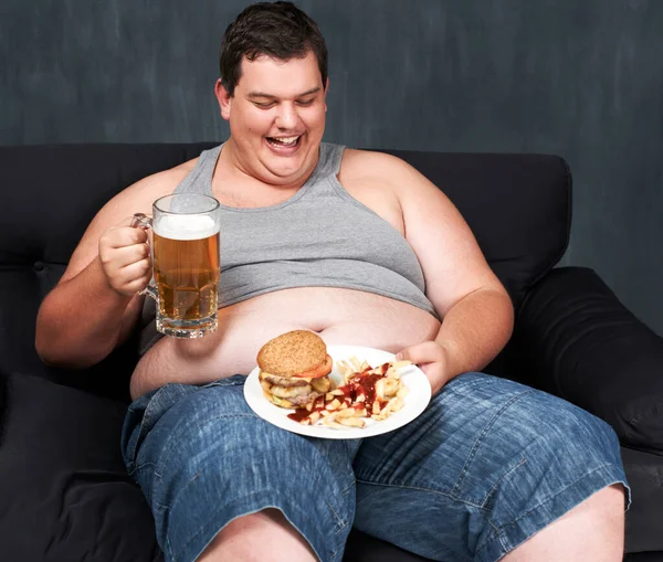 Algo para lavar su comida. Un joven obeso sentado en un sofá y hurgando en cerveza y comida rápida. —  Fotos de Stock