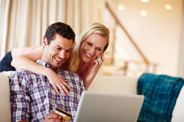 Einkaufen vom bequemen Sofa aus. Eine schöne Frau umarmt ihren Mann von hinten, während er auf der Couch sitzt und an seinem Laptop arbeitet. — Stockfoto