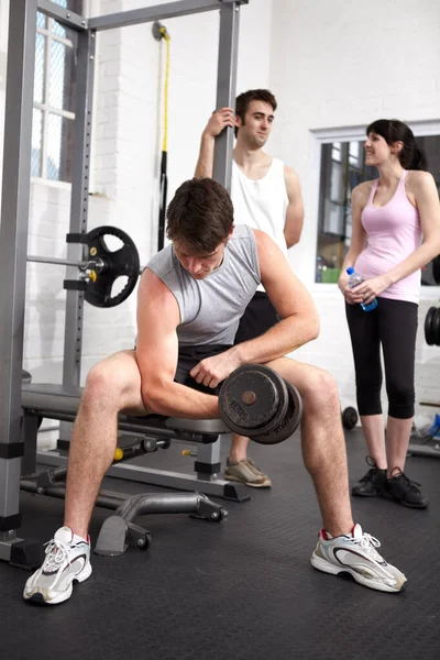 Mantenerse concentrado. joven culturista levantando un peso mientras dos personas charlan detrás de él en el gimnasio. — Foto de Stock