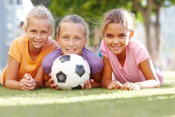 Nossa escola oferece meninas de futebol. Três garotas sorridentes deitadas em uma grama ensolarada posando com uma bola de futebol- copyspace. — Fotografia de Stock