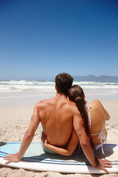 Everyting is so much more beautiful when youre inlove. Rearview shot of a young couple sitting on a surfboard while looking at the ocean. Stock Picture