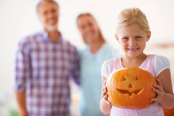 Orgogliosa della sua jack-o-lanterna. Ritratto di una bambina che tiene la sua jack-o-lanterna con i suoi genitori sfocati dietro di lei. — Foto Stock