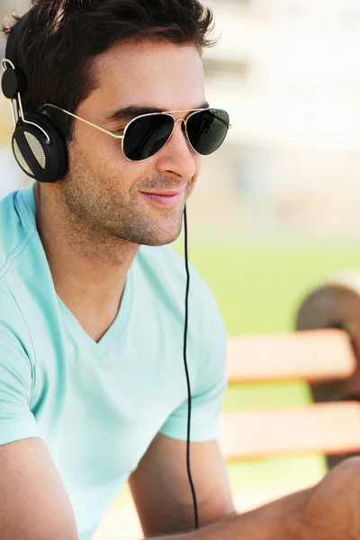 A afinar as suas músicas favoritas. Um jovem sorridente usando óculos de sol sentado em um banco em um parque e ouvindo música através de seus fones de ouvido. — Fotografia de Stock