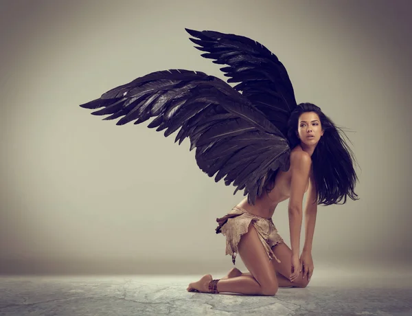 Dark angel of beauty. Shot of a woman with wings kneeling against gray background. Royalty Free Stock Photos