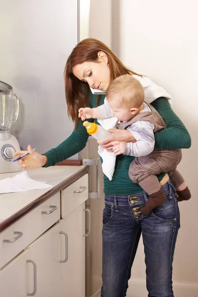 Mama zu sein, ist ein echter Jonglierakt. Junge Mutter versucht Multitasking, während sie ihren kleinen Sohn und seine Flasche hält. — Stockfoto