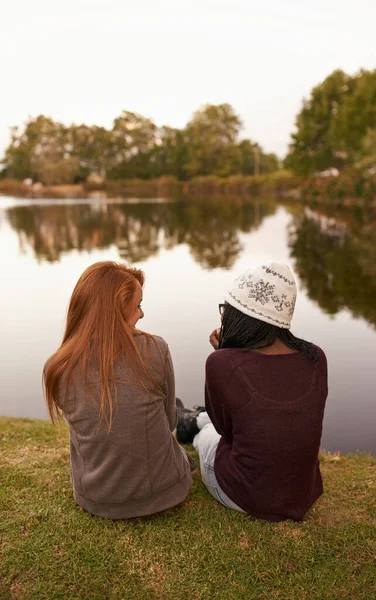 Qualitätszeit mit meinem Partner in der Kriminalität. Rückansicht von zwei Teenager-Mädchen, die neben einem See sitzen und plaudern. — Stockfoto