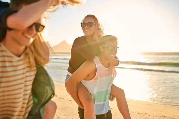 Szerelem, barátok és nevetés. Vágott lövés két szeretetteljes fiatal férfi piggyback barátnőik a strandon. — Stock Fotó