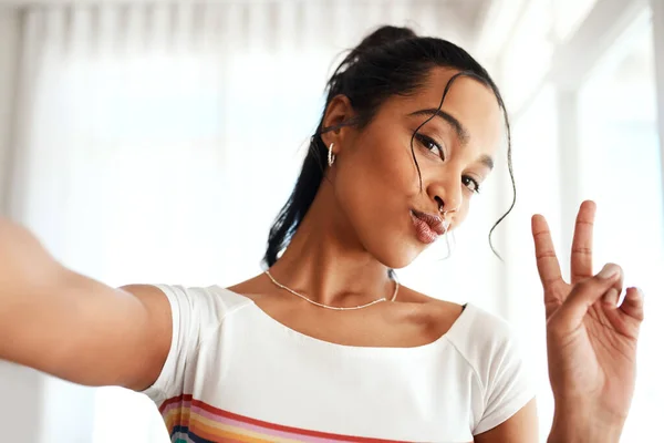 Dare a questo mondo una buona energia. Ritratto ritagliato di una giovane blogger attraente in piedi nella sua casa e fare un gesto di segno di pace per un selfie. — Foto Stock