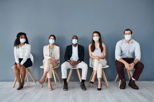 As precauções que tomarmos agora garantirão a nossa segurança e sucesso. Retrato de um grupo de empresários usando máscaras enquanto se sentam na fila contra um fundo cinza. — Fotografia de Stock