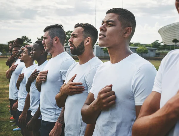 So weit sind wir. Schnappschuss eines Teams selbstbewusster junger Rugbyspieler, die vor einem Rugbyspiel auf einem Feld stehen und ihre Hymne singen. — Stockfoto