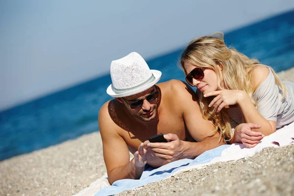 Staying in touch has never been easier. Shot of a young couple using a cellphone while suntanning on a beach. — Stock Photo, Image