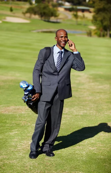 Doing business on the course. Shot of an african wearing a suit and talking on the phone while carrying a golf bag on a golf course. — Stock Photo, Image
