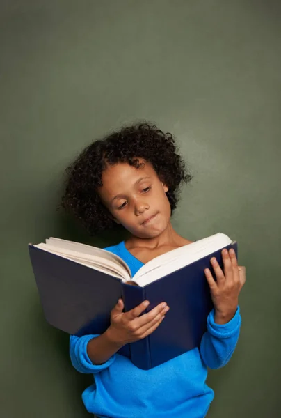 Er is geen vriend zo loyaal als een boek. Een jonge etnische jongen die een boek leest terwijl hij voor een schoolbord staat. — Stockfoto