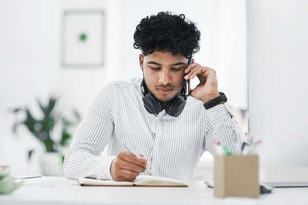 Can you repeat that please. Shot of a young businessman writing notes while talking on a cellphone in an office. — Stock Photo, Image