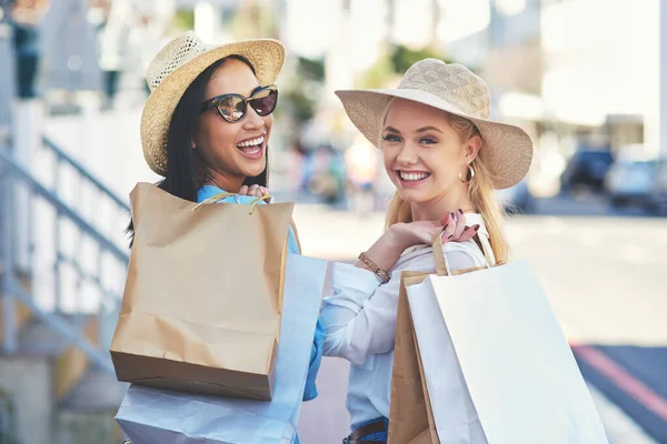 これは私たち自身を甘やかす私たちの小さな方法です。昼間に街のショッピングバッグと一緒に歩いている間に笑顔2人の魅力的な若いガールフレンドのトリミングされた肖像画. — ストック写真