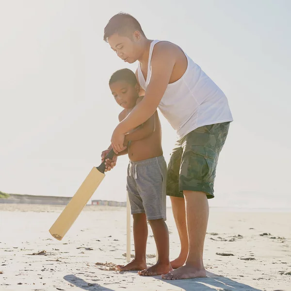 将来のクリケットスターを育てる。ビーチでクリケットバットを保持する方法を息子に示す父親のショット. — ストック写真