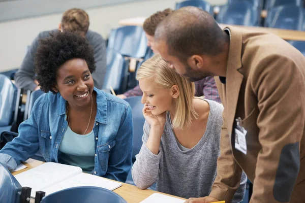 Een docent staat altijd voor je klaar als je vragen hebt. Een jonge man die met twee meisjes praat die in een collegezaal zitten.. — Stockfoto