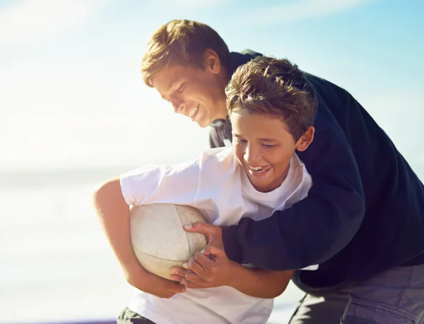 É só um jogo amigável entre irmãos. Tiro de dois irmãos felizes brincando com uma bola de rugby na praia. — Fotografia de Stock