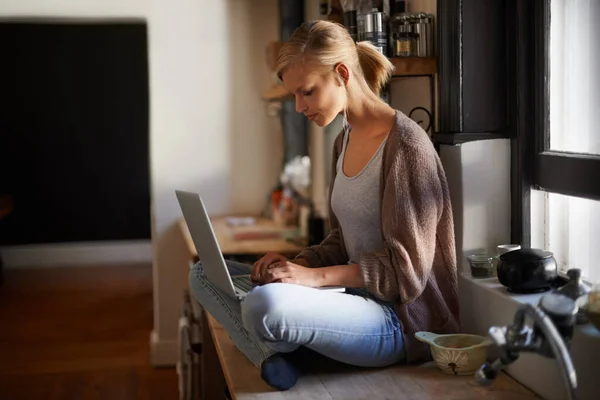 Ihre ganze Arbeit erledigt sie in der Küche. Eine attraktive junge Frau benutzt ihren Laptop, während sie auf ihrem Küchentisch sitzt. — Stockfoto