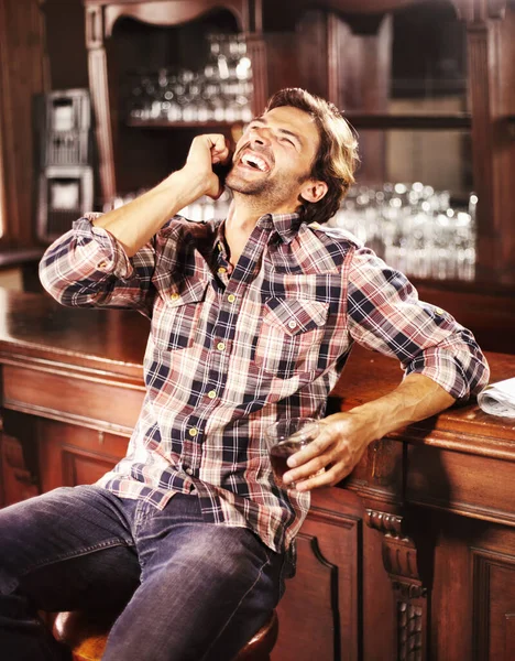 Das ist urkomisch. Schöner junger Kerl hockt mit einem Drink in der Hand an der Theke, während er mit seinem Handy telefoniert. — Stockfoto