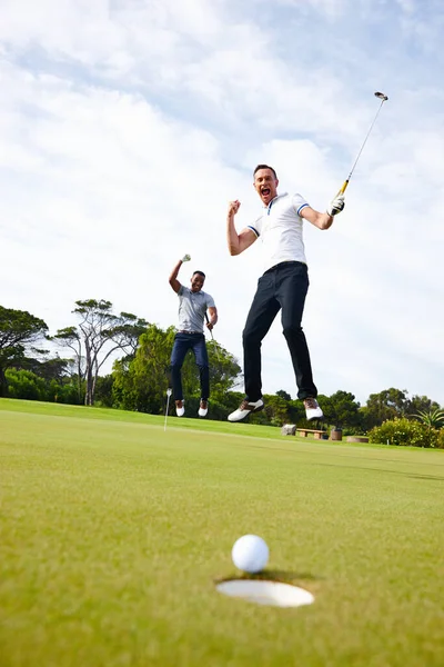 Πώς θα μπορούσε το γκολφ να είναι βαρετό; Χαμηλή γωνία βολή μιας μπάλας του γκολφ πλησιάζει την τρύπα, ενώ δύο παίκτες βλέπουν. — Φωτογραφία Αρχείου
