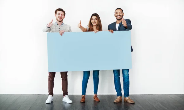 Machen Sie mit. Studioaufnahme einer Gruppe junger Leute, die ein leeres Plakat in der Hand halten und Daumen nach oben vor weißem Hintergrund zeigen. — Stockfoto