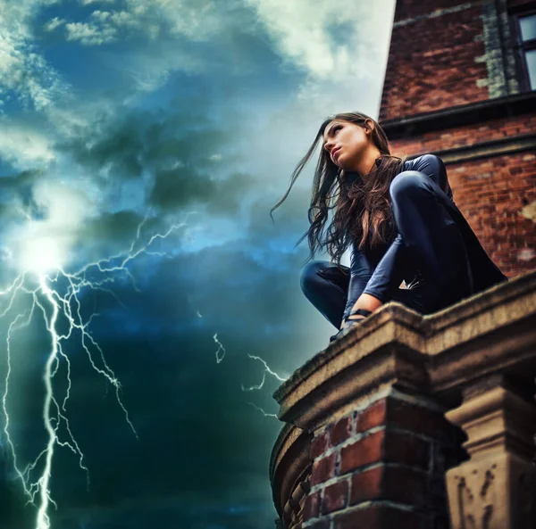 Ella es una fuerza de la naturaleza - Superhéroe. Mujer joven seductora en un catsuit negro agachado en una azotea en una tormenta relámpago. —  Fotos de Stock