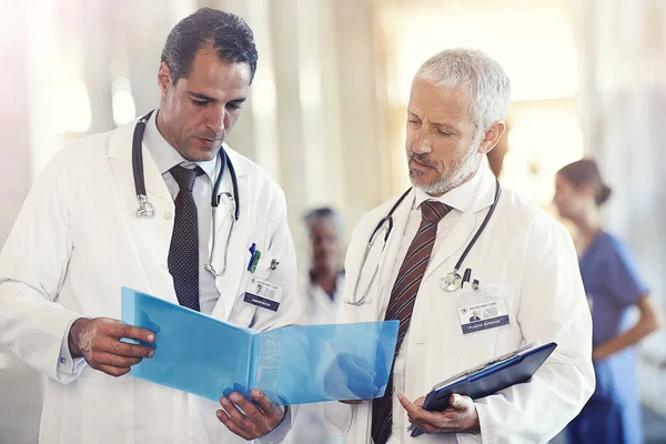 Necesito su consejo, doctor... Fotografía de dos médicos mirando un historial de pacientes con colegas borrosa en el fondo. — Foto de Stock