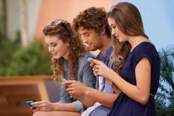 Ninguno de esos incómodos momentos de silencio aquí... Tres adultos jóvenes usando sus teléfonos celulares mientras están sentados al aire libre. — Foto de Stock