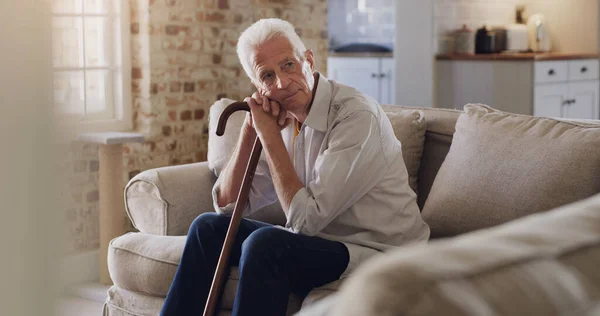 Where did all the time go. Shot of a senior man sitting alone on the sofa at home and looking contemplative while holding his walking stick. — Stock Photo, Image