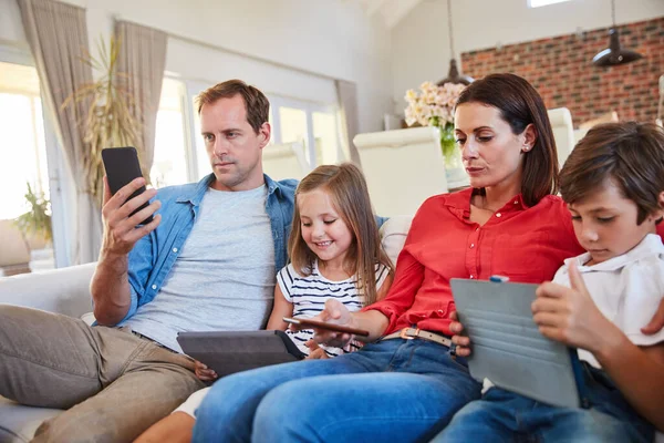 Distraciones digitales. Fotografía de una joven familia sentada junta en su sofá de la sala de estar distraída por varios medios y dispositivos. — Foto de Stock