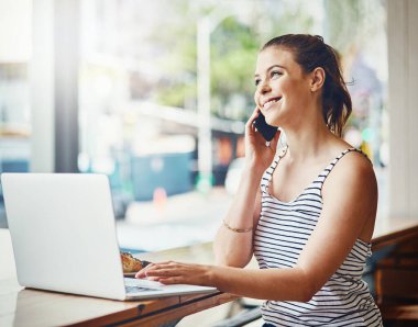 Rahatlatıcı bir atmosferde üretken kalmak. Cep telefonuyla konuşan ve kahve dükkanında laptopunu kullanan genç bir kadının fotoğrafı..
