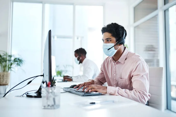 Tornare a fare affari. Foto di un giovane agente di call center che indossa una maschera facciale mentre lavora su un computer in un ufficio. — Foto Stock