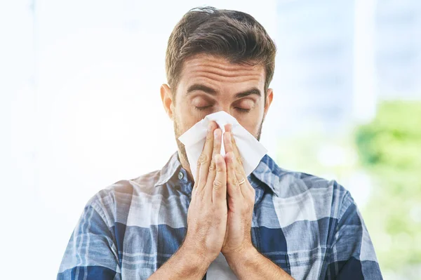 Going t have to stock up on tissues. Portrait of an uncomfortable looking young man blowing his nose with a tissue inside of a building during the day. — Stock Photo, Image