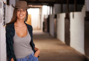 Just horsin around. A portrait of a beautiful young cowgirl leaning against a wall in a stable. clipart