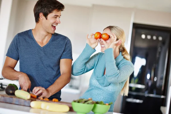 Verrückt spielen in der Küche. Aufnahme einer verspielten jungen Frau, die lustige Gesichter mit Gemüse macht, während ihr Mann eine Mahlzeit zubereitet. — Stockfoto