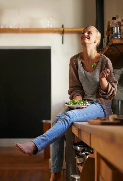 她很喜欢她的新饮食。拍了一个漂亮的年轻女人坐在厨房柜台边吃沙拉边笑. — 图库照片
