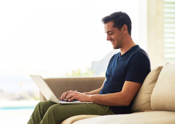 Pasar algo de su bien ganado tiempo libre en línea. El disparo de un joven guapo usando su portátil mientras estaba sentado en el sofá en casa. — Foto de Stock