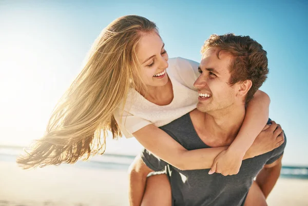 Du gör den här underbara dagen ännu vackrare. Skjuten av ett lyckligt ungt par njuter av en piggyback rida på stranden. — Stockfoto