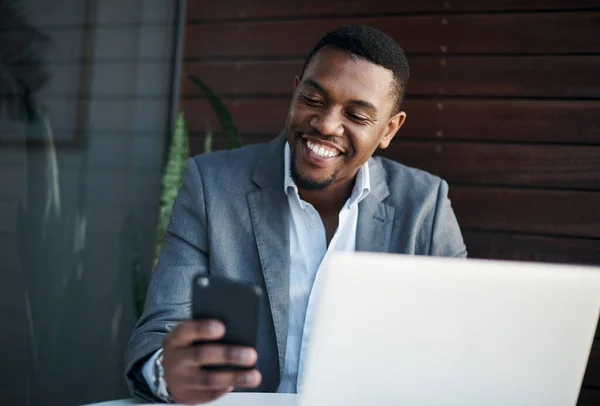 Clientes felices es igual a un gran negocio. Foto de un joven hombre de negocios guapo sentado solo en la oficina mientras usa su teléfono celular y su computadora portátil. —  Fotos de Stock
