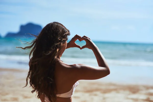 Adoro a praia. Tiro de uma jovem mulher moldando um coração enquanto olha para a paisagem na praia. — Fotografia de Stock
