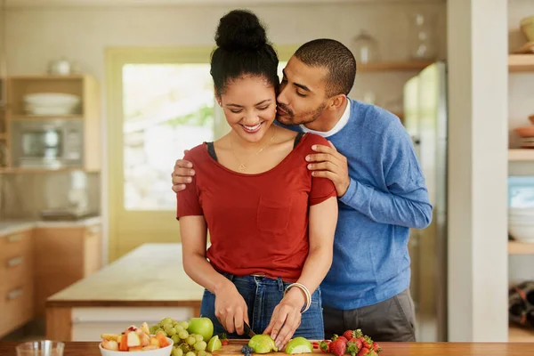Apoiando-se mutuamente em ser o mais saudável que podem ser. Tiro de um jovem beijando sua esposa enquanto ela prepara um lanche saudável em casa. — Fotografia de Stock